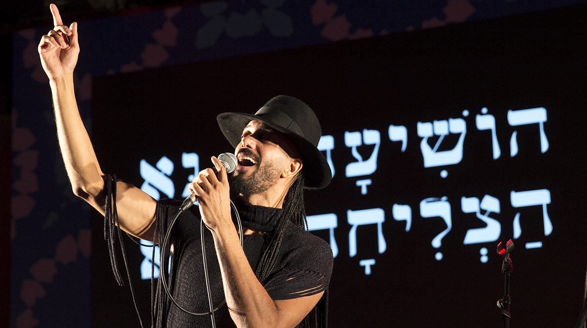 Frontman Ravid Kahalani points upwards as he sings on stage, against Hebrew writing in the backdrop.